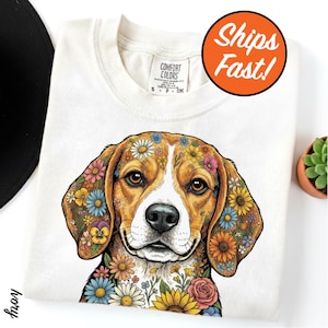 May include: Ivory-colored sweatshirt featuring a detailed illustration of a beagle with colorful floral patterns on its fur. The dog's eyes are brown, and the flowers include daisies, roses, and pansies. The shirt has a crew neck and a Comfort Colors tag.