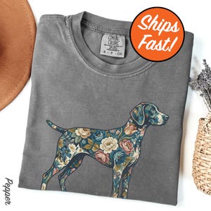 May include: Gray Comfort Colors t-shirt featuring a floral patterned dog design. The dog is adorned with blue, pink, and cream-colored flowers. The shirt has a crew neck and short sleeves. The tag reads "Comfort Colors".