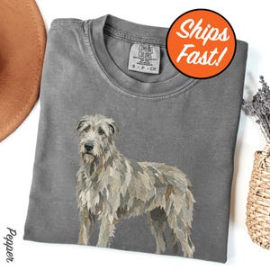 May include: A gray Comfort Colors t-shirt featuring a detailed illustration of an Irish Wolfhound. The dog is depicted in shades of gray and beige. The shirt has a crew neck and a small tag. An orange circle with the words "Ships Fast!" is visible.