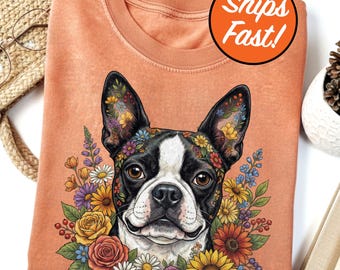 May include: A coral-colored sweatshirt featuring a Boston Terrier with a floral design. The dog has black and white markings and is surrounded by colorful flowers. The image includes the text "Ships Fast!" in an orange circle.