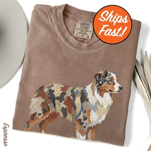 May include: A brown Comfort Colors t-shirt featuring a colorful collage-style image of an Australian Shepherd dog. The dog's fur is depicted with various paper scraps. The shirt has a crew neck and a relaxed fit.