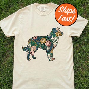May include: Cream-colored t-shirt featuring a floral patterned dog silhouette. The dog design incorporates various flowers in shades of pink, blue, and yellow against a green background. The shirt has a classic crew neck and short sleeves.