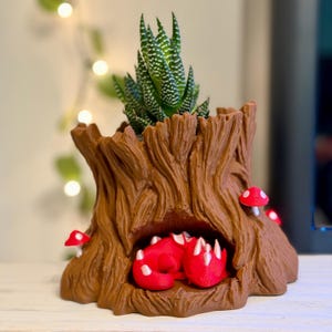 May include: A brown tree stump planter with a textured bark finish, featuring a small red dragon figurine resting inside a hollow cavity. The planter is decorated with small red mushrooms with white spots and contains a green succulent plant.