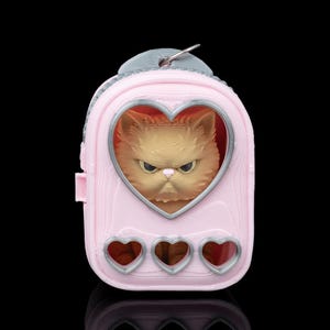 May include: A light pink, heart-shaped pet carrier keyring featuring a 3D-printed orange cat with a grumpy expression visible through a clear window. The carrier has a silver-coloured metal ring at the top and three small heart-shaped cutouts at the base.