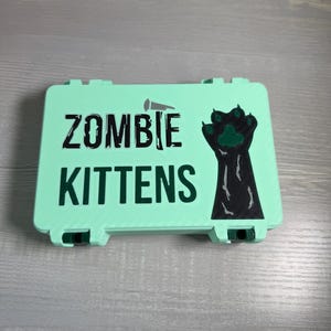 May include: A mint green rectangular case with the words "ZOMBIE KITTENS" in black, stylized lettering. A black paw print with green claws is on the right side. The case has a textured surface and black hinges.