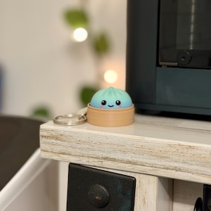 May include: A small, round, light blue and teal dumpling-shaped figurine with a smiling face, black eyes, and pink cheeks, resting inside a tan, cylindrical, basket-like container. A silver metal keychain ring is attached to the side of the base.