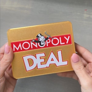 May include: A gold-colored Monopoly Deal card game box. The box features the Monopoly logo with the iconic top-hatted character and the word "MONOPOLY" in red. The word "DEAL" is prominently displayed in white.