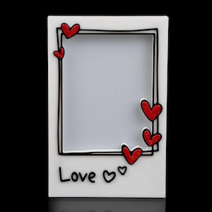 Puede incluir: Un marco de fotos blanco con borde negro y detalles de corazones rojos. La palabra "Love" está escrita en negro en la parte inferior, con dos símbolos de corazón. El marco está diseñado para una fotografía.
