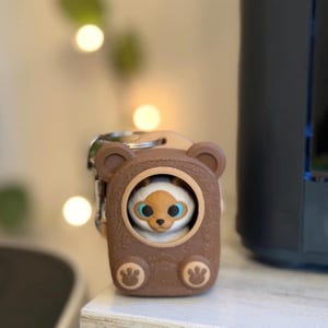 May include: A brown bear-shaped keychain case featuring a circular cutout that reveals a small, white, cat-like figure with blue eyes and brown ears. The case has textured wood-grain detailing and two small paw prints at the bottom.