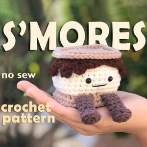 May include: A crocheted s'more plush toy with a brown chocolate top, white marshmallow middle, and brown graham cracker bottom. The toy has a smiling face with black eyes and brown crocheted hair. The toy is sitting in a hand, and the text "S'mores no sew crochet pattern" is visible in the background.