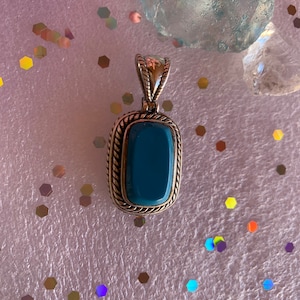 May include: A silver pendant with a blue gemstone set in a square frame. The pendant has a rope-like border around the frame.