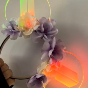 May include: A headband with purple and white flowers and two iridescent circles with the letters "BTS" inside. The circles are lit up with a warm orange glow.