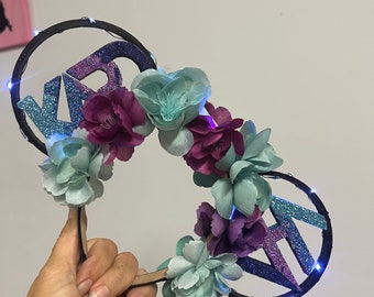 May include: A black headband with two circular light-up ears featuring the letters "KR" in glittery purple and blue. The headband is decorated with light blue and purple fabric flowers.