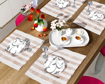 May include: A wooden table with a white and brown striped placemat, a white plate with a gray floral design, and a clear glass with a stem. There are also flowers in a white vase and a tray with cups and saucers.