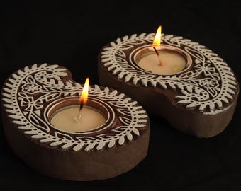 May include: Two brown, paisley-shaped candle holders with white floral designs. Each holder contains a lit tea light candle. The candle holders are made of wood and sit on a black surface. The flames are bright orange.