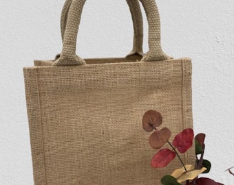 May include: A small, brown burlap tote bag with double handles. The bag is sitting on a white surface with a sprig of dried leaves in the corner.