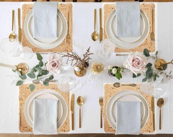 May include: Four place settings with woven placemats, white plates, light blue napkins, gold silverware, and clear glasses. The table is set with white flowers and greenery.