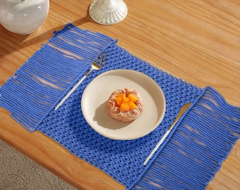 May include: A blue woven placemat with fringe edges. The placemat is set on a wooden table with a white plate and a fork and knife.