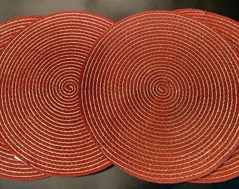 May include: Set of five round placemats in a warm reddish-brown hue. Each mat features a spiral design with white concentric lines radiating from the center. The placemats are made of a textured material.