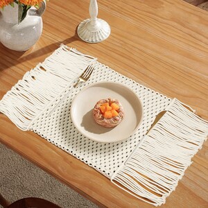 May include: A white crocheted placemat with fringe on all sides. The placemat is on a wooden table with a white plate and a small pastry on top.
