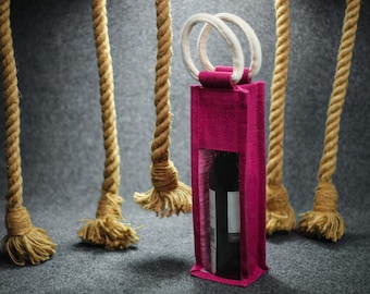 May include: A pink jute wine bottle carrier with a clear plastic window, showing a bottle of wine inside. The carrier has a white rope handle and is surrounded by brown ropes.