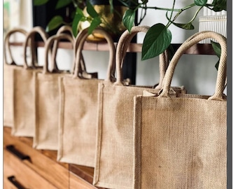 May include: A set of six brown burlap tote bags with rope handles. The bags are lined up on a wooden surface.