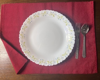 May include: A white dinner plate with a floral design sits on a red placemat with a fork and spoon to the right.