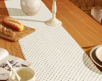May include: A white crocheted table runner with a diamond pattern. The runner is on a wooden table with a vase of orange flowers, a candle holder, and a book.