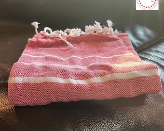 May include: Red and white striped Turkish towel with fringe. The towel is folded and lying on a brown leather surface.
