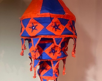 May include: A multi-tiered, hanging lampshade made of fabric. The lampshade is orange and blue with a geometric pattern and star-shaped embellishments. The lampshade has tassels hanging from the bottom.