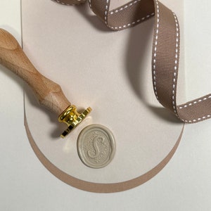 May include: A wooden handle with a gold metal stamp and a beige wax seal with a decorative letter S. The stamp is on a beige surface with a brown ribbon with white stitching.
