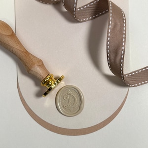 May include: A wooden handle wax stamp with a gold metal base and a beige wax seal with a decorative letter D design.