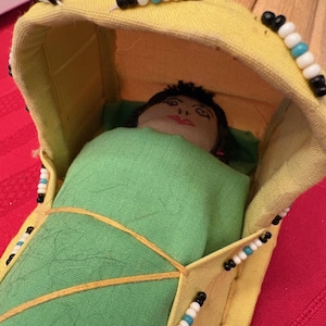 May include: A handmade doll in a yellow and tan cradle. The cradle is decorated with black, white, and blue beads. The doll is wrapped in a green blanket and has a painted face.