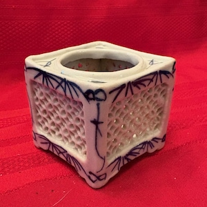 May include: A square, white ceramic container with a circular opening and intricate lattice-like cutouts. The edges are adorned with blue, stylized floral and geometric patterns. The container sits on a red surface.