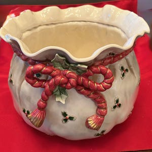 May include: A ceramic planter with a ruffled rim and a cream-colored interior. The exterior is decorated with a red rope bow and holly accents. The planter is set on a red surface.