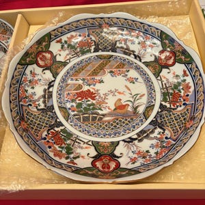 May include: A decorative ceramic plate with a scalloped edge. The plate features a detailed floral and bird design in red, orange, blue, and green. The plate is in a yellow box, suggesting it is part of a set.