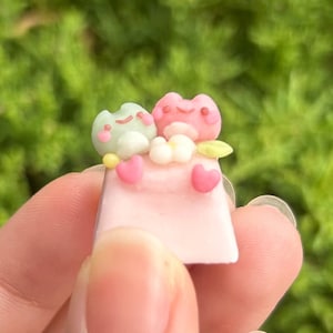 Könnte beinhalten: Zwei kleine Froschfiguren, eine grün und eine rosa, sitzen auf einer rosa Keycap mit weißen Blumen und Herzen. Die Keycap wird von einer Hand gehalten.