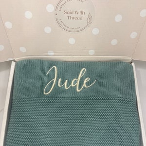 May include: A teal knitted blanket, embroidered with the name "Jude" in cream script, presented in an open white box. The box has a polka-dot interior and a circular sticker with the text "Bespoke Embroidery Said With Thread & Personalisation".