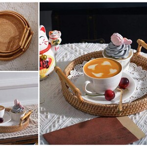 Może przedstawiać: Okrągłe plecione tace z dwoma bambusowymi uchwytami, na którym znajduje się filiżanka kawy z sercowatym wzorem latte art, babeczka z różowym lukrem i czerwone wiśnie. Tace znajduje się na białym obrusie z koronkową serwetką.