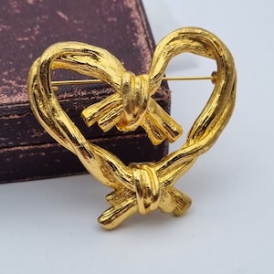 May include: A gold-toned heart-shaped brooch with a knotted design. The brooch is made of twisted metal and has a pin on the back.
