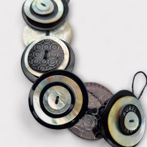 Può includere: Un braccialetto di bottoni bianco e nero con un motivo circolare. Il braccialetto è composto da più strati di bottoni, ognuno con un design diverso. Il braccialetto ha un cordoncino nero con un anello all'estremità. Sul braccialetto è scritta la parola "AUREL" su uno dei bottoni.