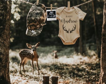 May include: A baby onesie with the text "Baby Andrew" and a pair of antlers printed on it. The onesie is hanging on a clothesline in a woodland setting with a deer in the background. A camouflage hat and a sonogram picture are also hanging on the clothesline.