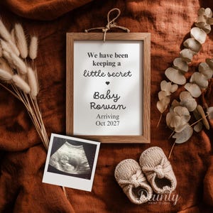 May include: A wooden frame containing a sign that reads We have been keeping a little secret Baby Rowan Arriving Oct 2027, accompanied by a sonogram photo and a pair of small beige knit baby booties, all arranged on a textured rust-colored fabric.