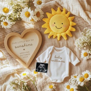 May include: A heart-shaped wooden frame with the text "OUR LITTLE RAY OF sunshine ARRIVING OCTOBER 202X" is next to a white baby onesie with the name "baby williams" in gold script. A yellow knitted sun and ultrasound image are also present.