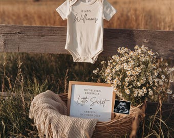 May include: Cream-colored baby onesie with the name "Williams" printed on it, hanging on a clothesline. A wooden fence and tall grass are in the background. A basket holds a blanket, flowers, a sign that reads "We've been keeping a Little Secret" and a sonogram.