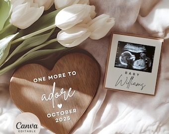 Puede incluir: Un letrero de madera en forma de corazón con el texto "One More To Adore" y "October 2025". Un pequeño corazón blanco se encuentra entre las palabras "adore" y "October". Una imagen de ultrasonido enmarcada con el texto "Baby Williams" está al lado del letrero.