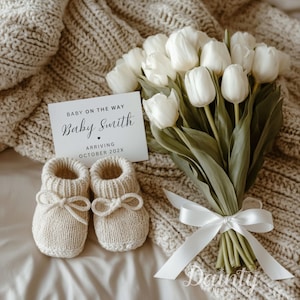 May include: A baby announcement card with the text "Baby on the Way" and "Baby Smith" is displayed next to a pair of knitted baby booties and a bouquet of white tulips. The card indicates an October 202X arrival. The scene is set on a neutral-toned blanket.