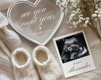 Puede incluir: Un espejo en forma de corazón con el texto "see you next year" y "January 202X". Junto a él, un par de patucos de bebé color crema y una ecografía con el nombre "Alexander". Flores blancas y tela suave completan la escena.