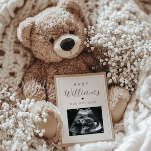 May include: A plush brown teddy bear rests on a cream-colored blanket, surrounded by white flowers. A card with the text "BABY Williams DUE OCT 202X" and an ultrasound image is in front of the bear, announcing a baby's arrival.