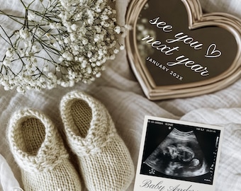 Puede incluir: Un anuncio de bebé con patucos de punto, un espejo en forma de corazón con la frase "see you next year" y una imagen de ultrasonido con "Baby Anderson". Flores blancas y tela suave completan la escena.